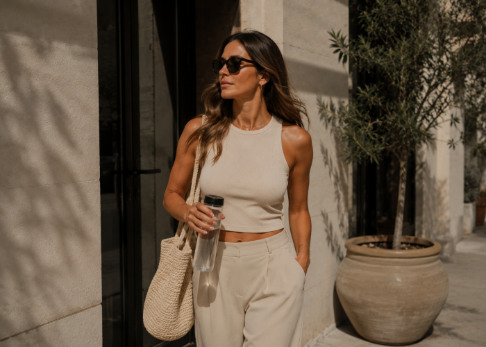 Woman walking outdoors in neutral-toned clothing with sunglasses and a water bottle in warm natural sunlight.
