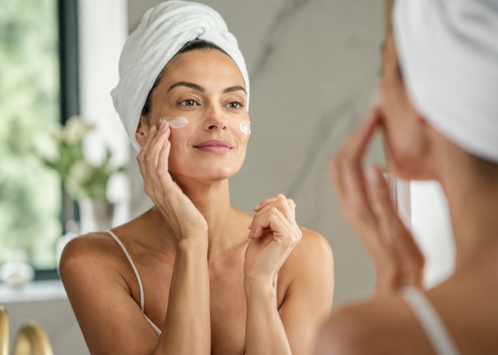 Woman applying facial skincare cream while looking into a bathroom mirror with a towel wrapped around her hair.