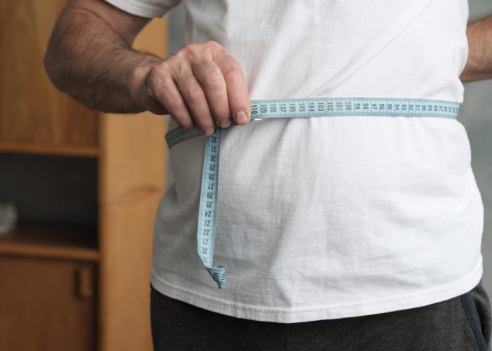 Fat mature man measuring his belly with measurement tape standing in the living room. Problem with overweight, bad health and dieting concept